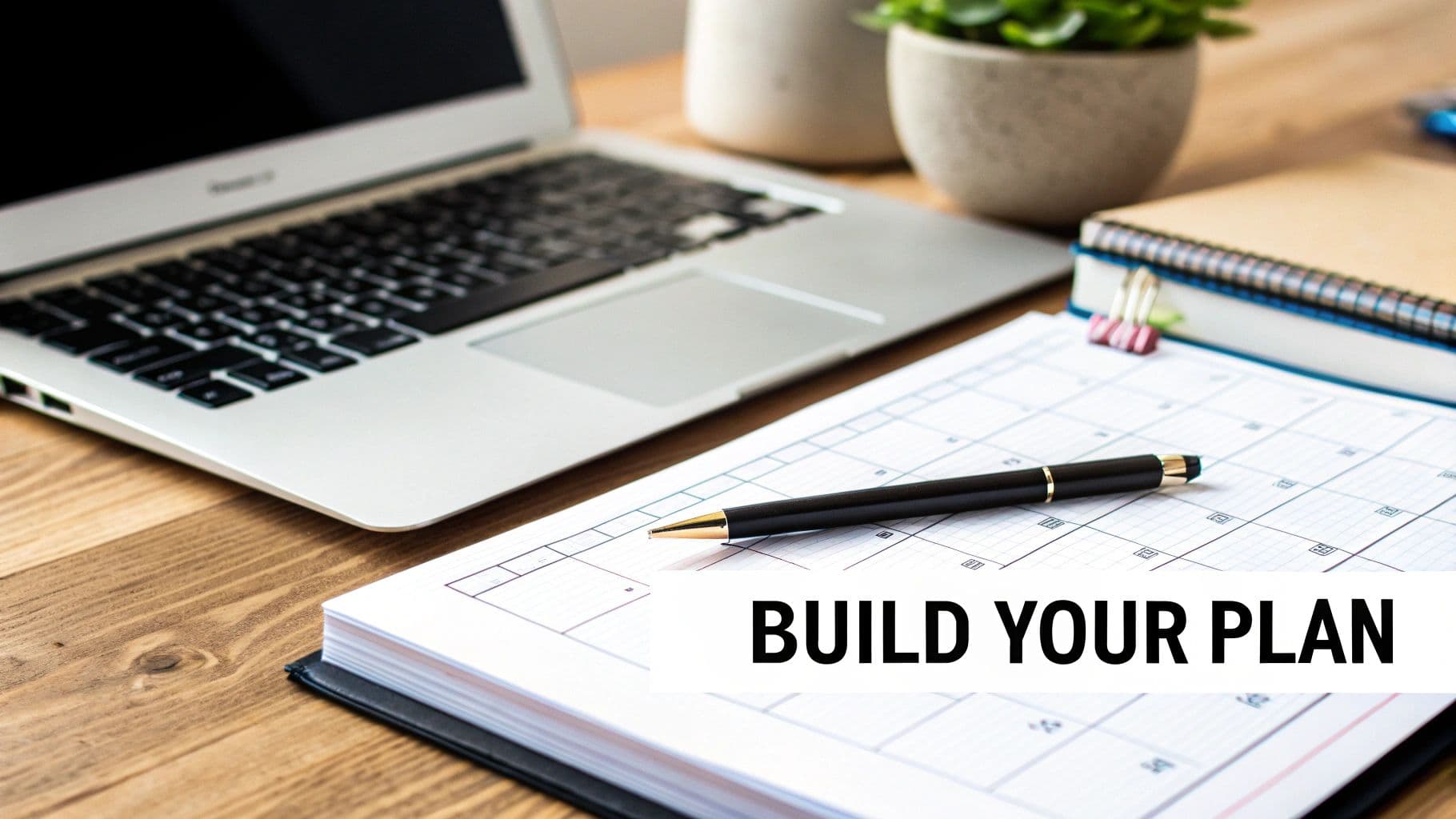 A laptop, green plant, spiral notebook, and a pen on a planner displaying 'BUILD YOUR PLAN' on a wooden desk.